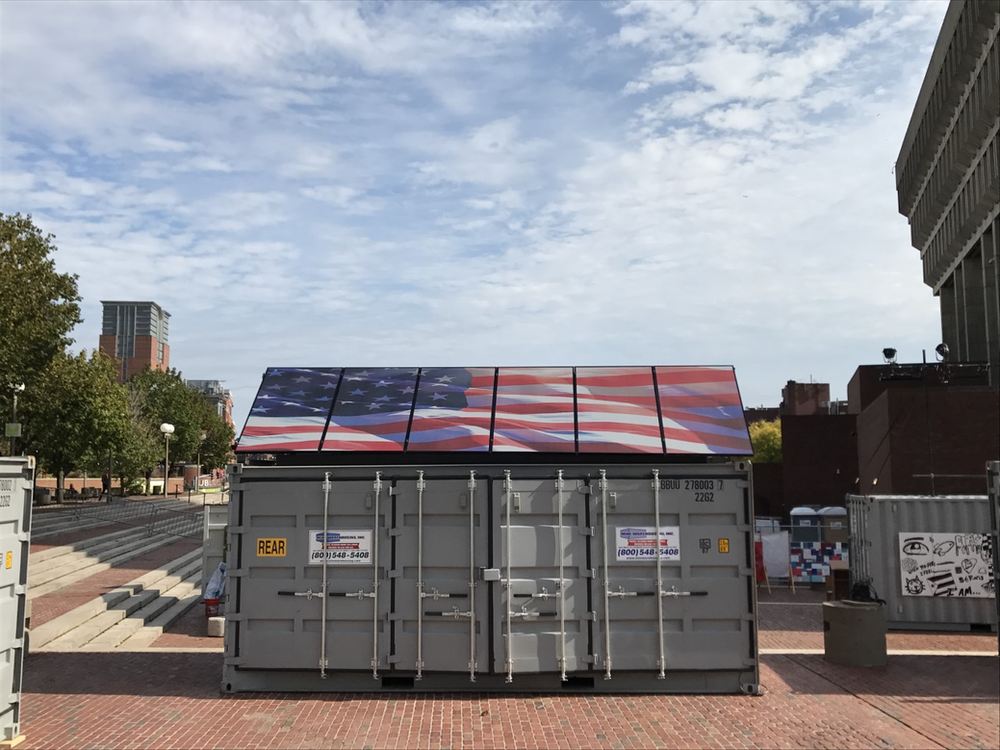 Photo-realistic graphic of American flag billowing in wind angle on top of solar panels, angled 45 degrees atop gray cargo box in a town square in Boston