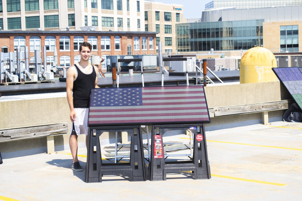Stiched design of American flag on rectangular solar panel, exposed to sunshine on a rooftop in the middle of sunny day. Decorated solar panel sits atop two black benches with VP of Product holding the panel.
